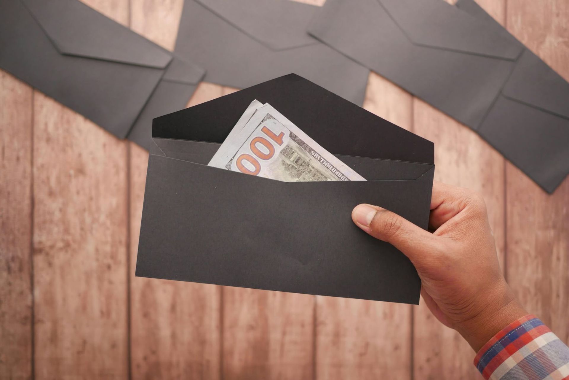 Mão segurando um envelope preto com dinheiro e outros envelopes sobre uma superfície de madeira.