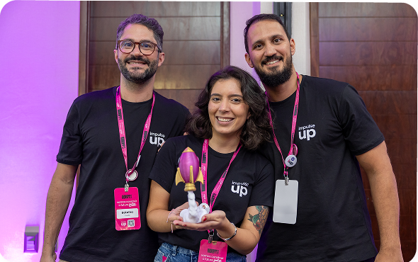 Três pessoas vestindo camisas pretas, segurando um troféu e sorrindo, têm como pano de fundo um cenário de evento.