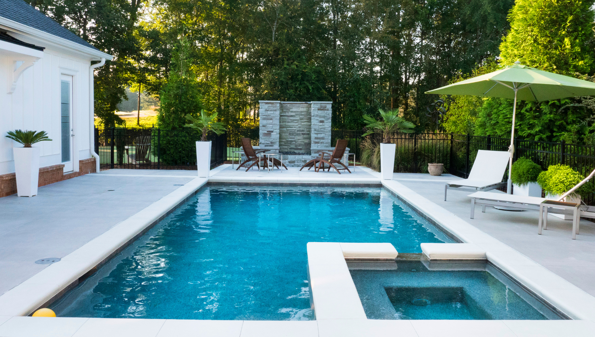 Rectangular swimming pool and hot tub with lounge chairs, umbrella, and a water feature.