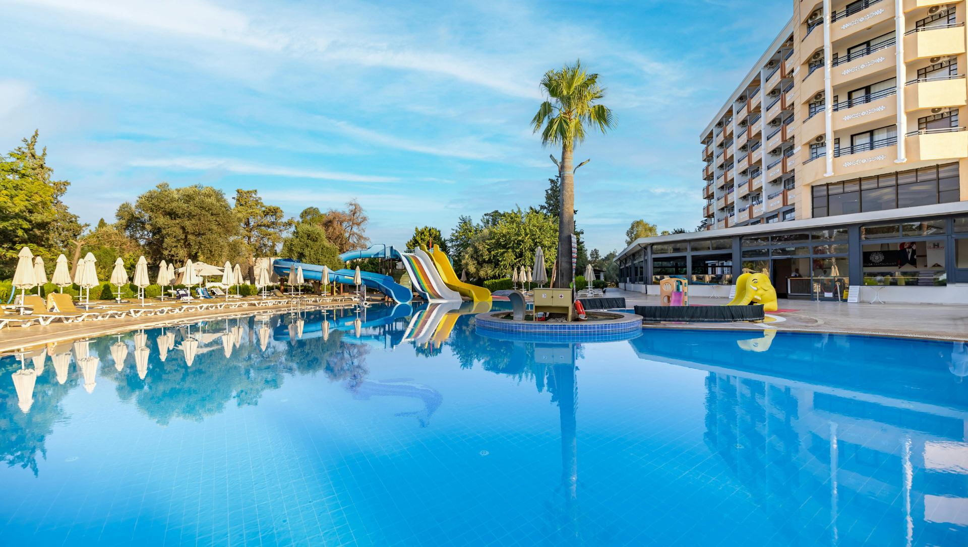 Swimming pool at a resort with palm trees, water slides, and hotel building. Sunny day.