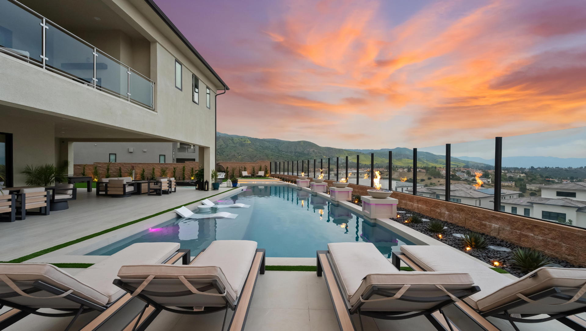 Luxury home with long pool overlooking city at sunset. Beige loungers face the water.