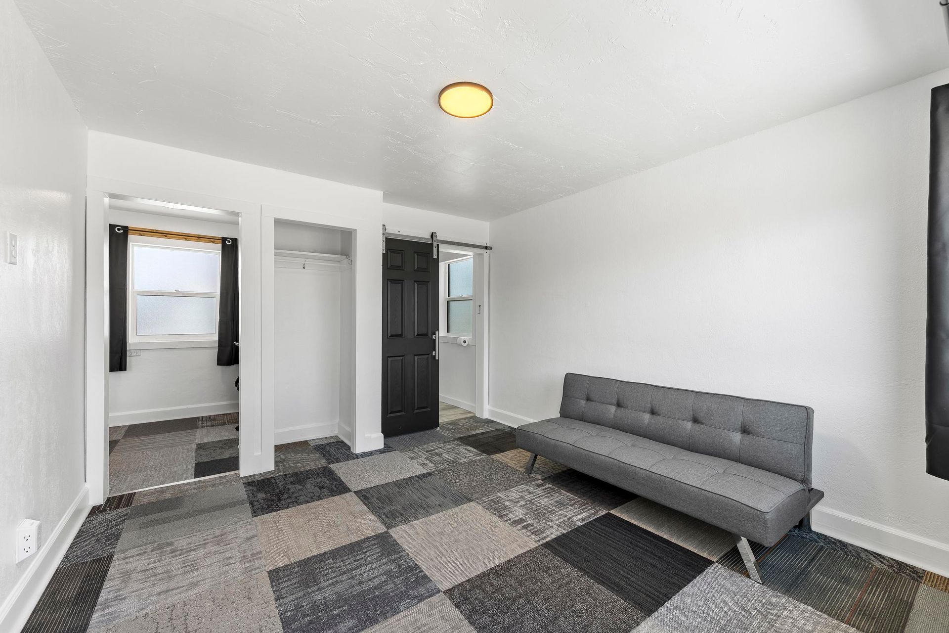 Bright bedroom with gray sofa bed, patterned rug, white walls, closet, and bathroom doorway.