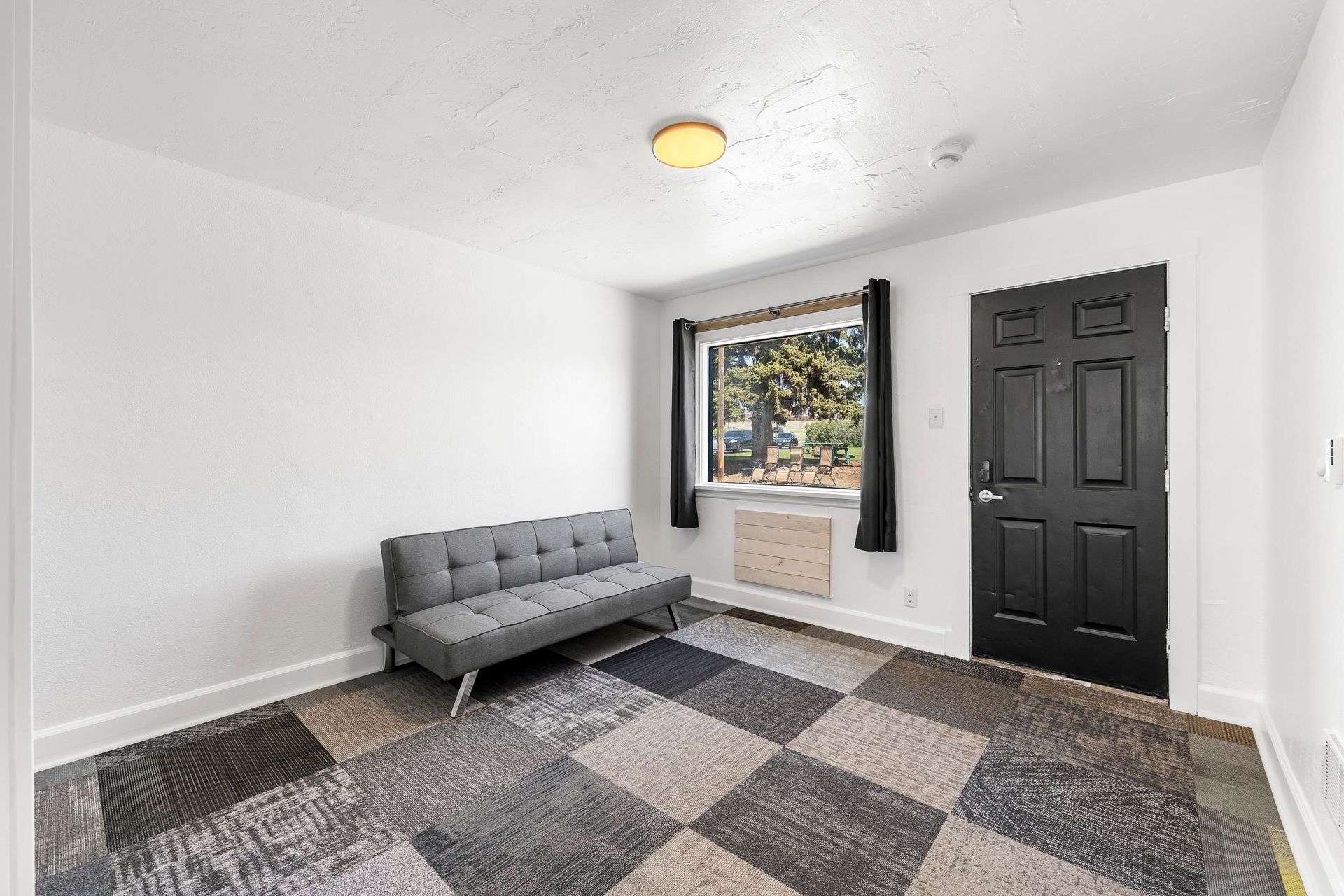 Small white room with a gray sofa, black door, window, and checkered carpet.