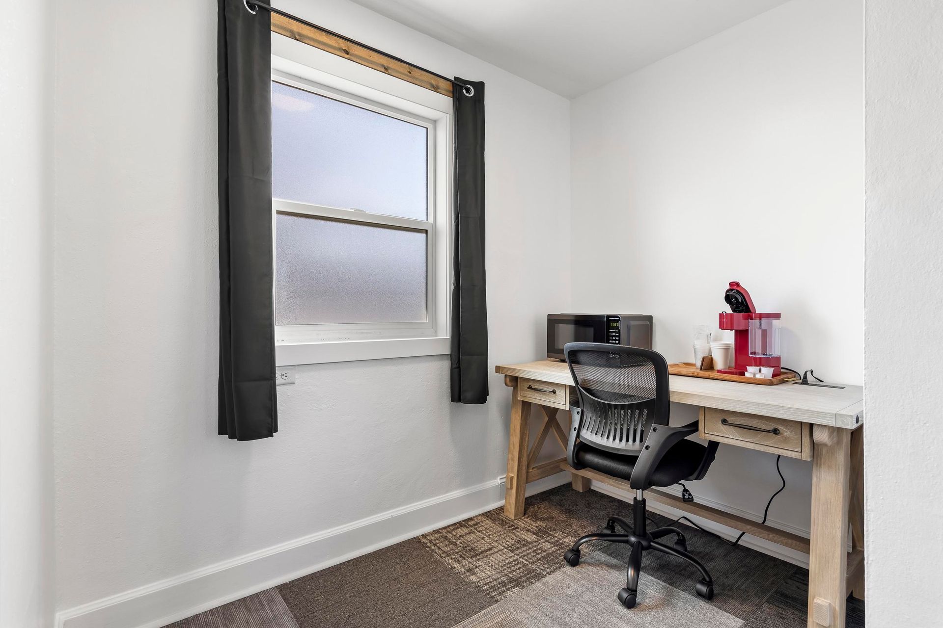 Small home office with a desk, chair, and window with dark curtains on white walls.