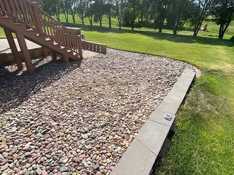Brown wooden stairs next to a gravel bed, bordered by a low stone wall and green grass.