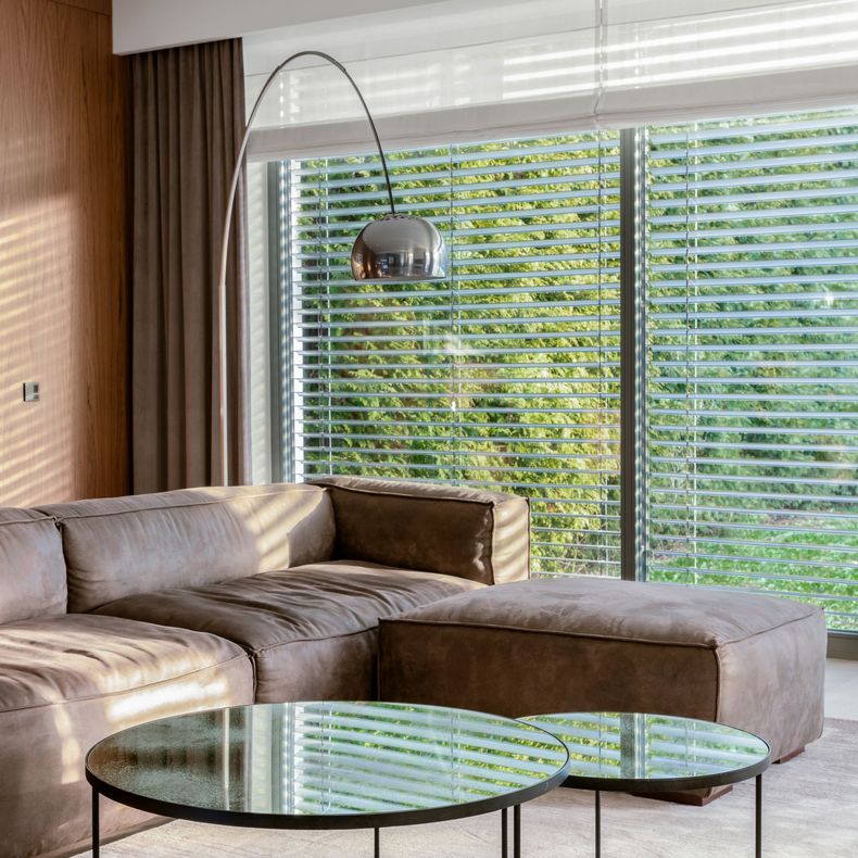 Modern living room with brown sectional sofa, two glass coffee tables, and arched lamp. Large window with blinds.