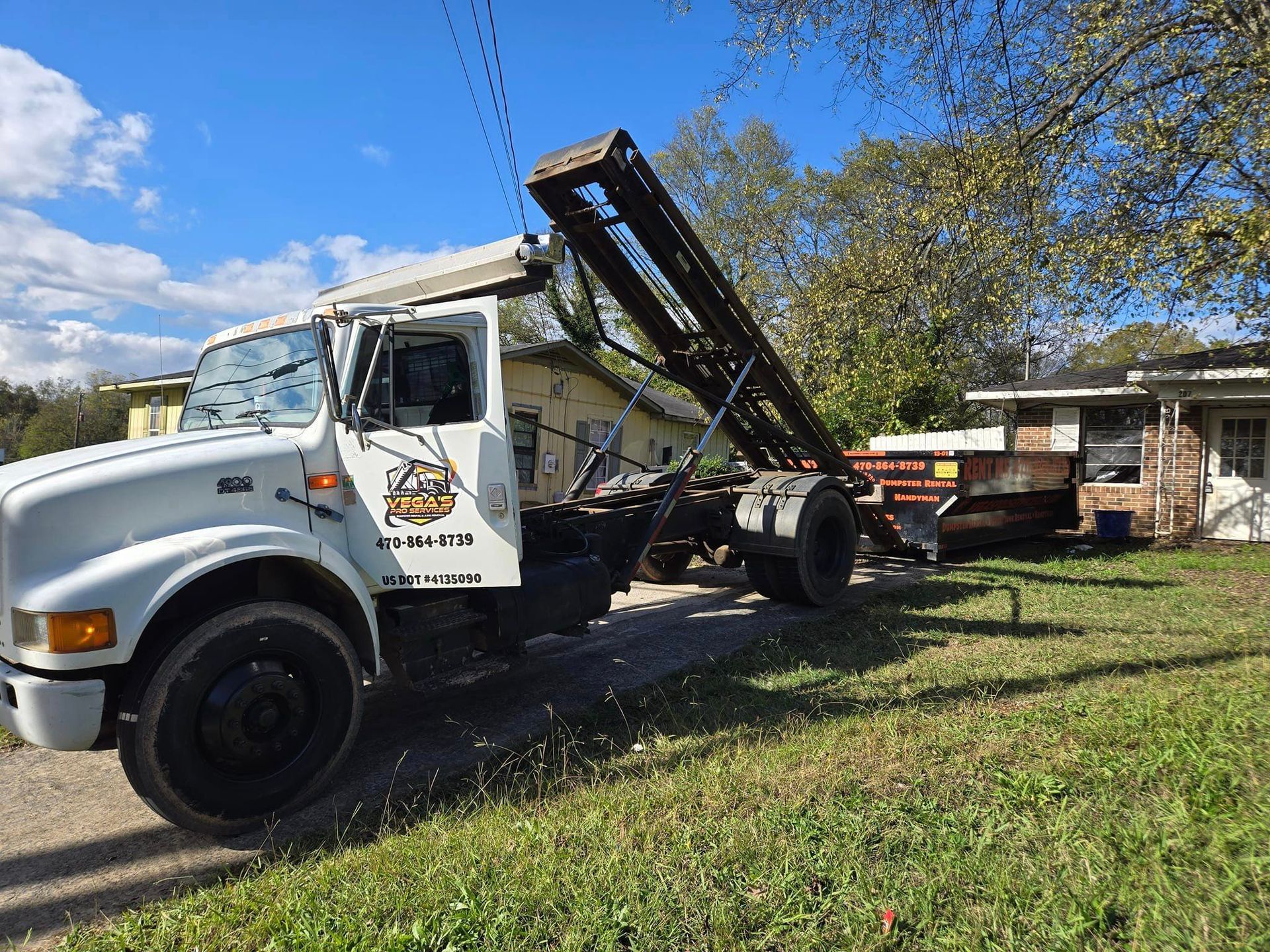 White roll-off truck with raised bed, parked in front of a house on a sunny day.