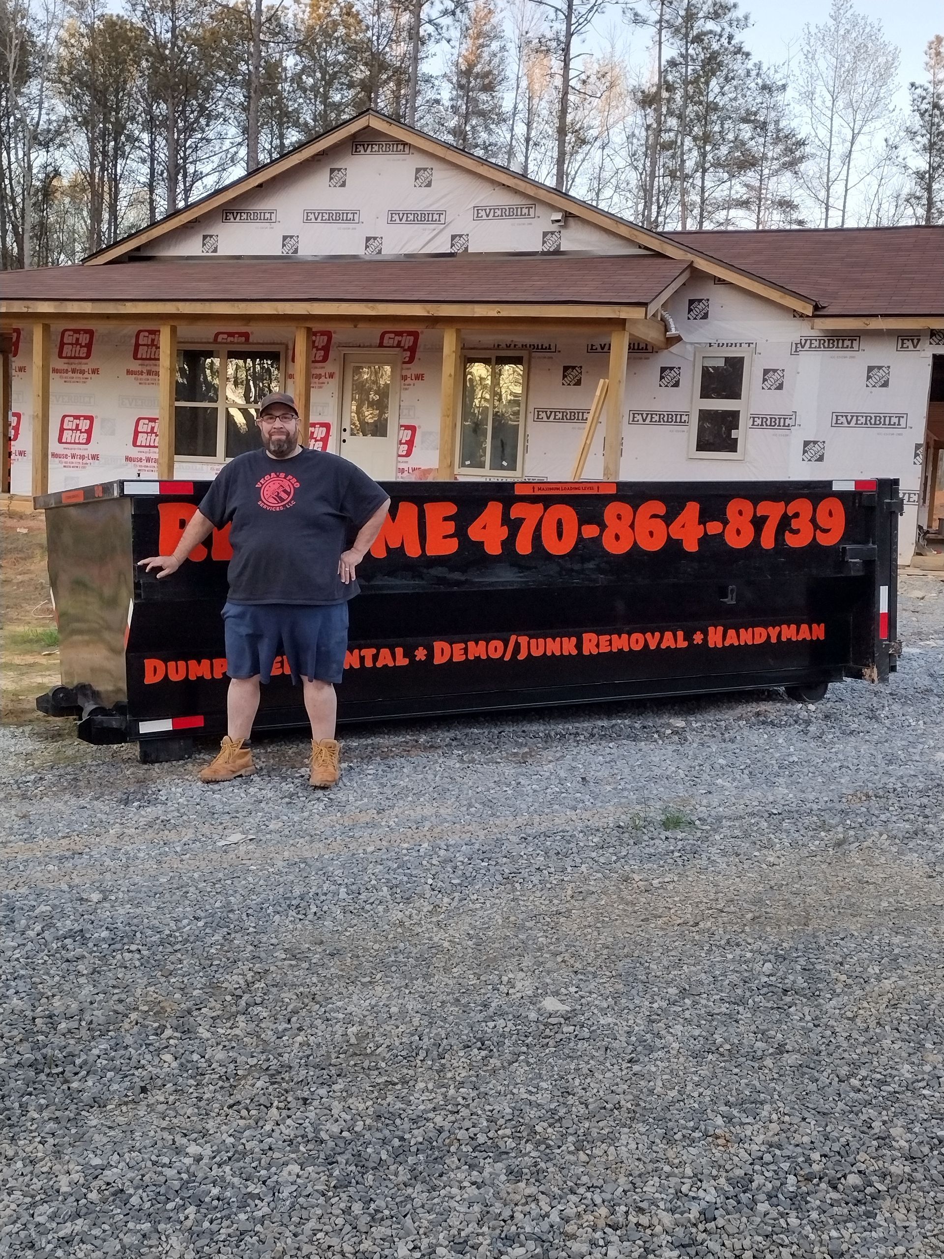 Man stands next to a dumpster in front of a house under construction. Dumpster has a phone number.