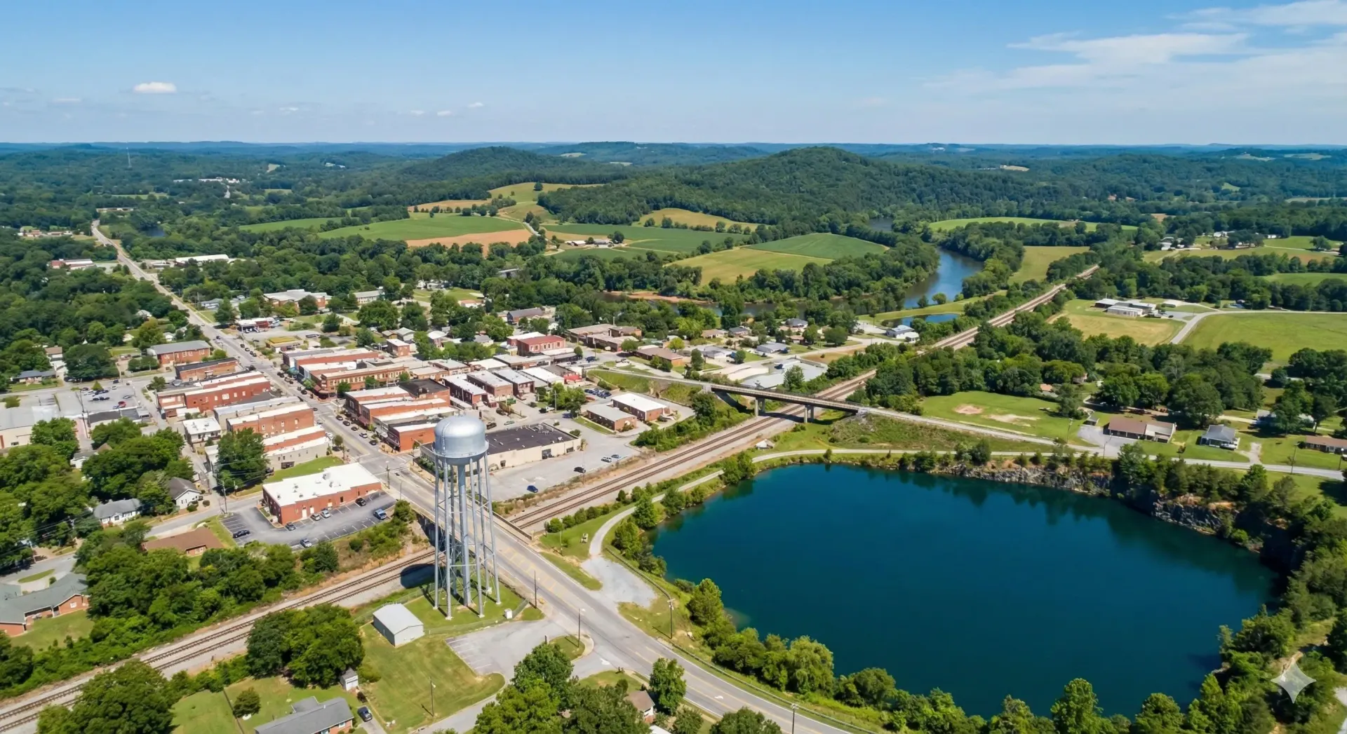 Aerial view of a town with buildings, a lake, railroad tracks, and surrounding green hills under a blue sky.