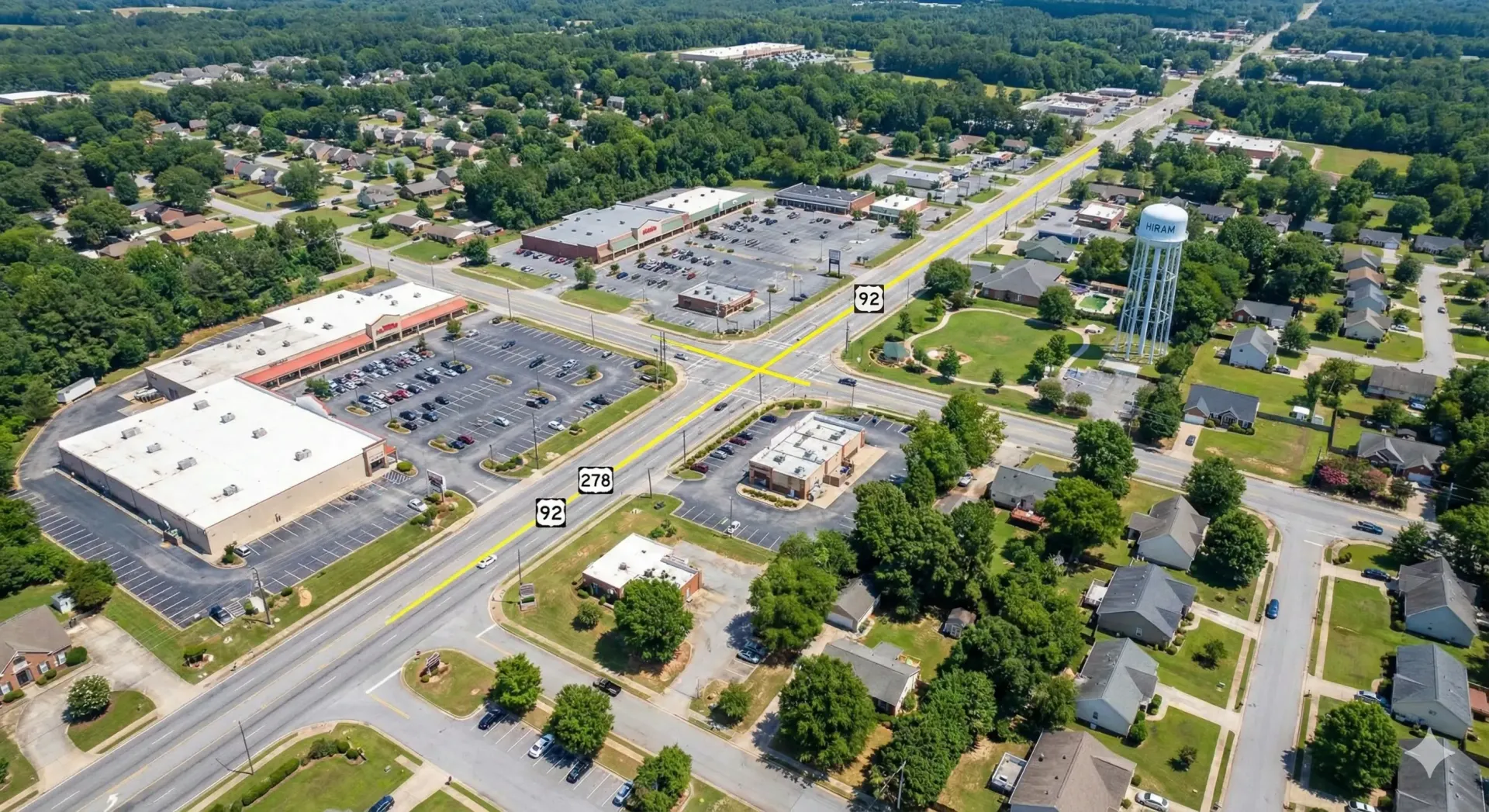 Aerial view of a town intersection with commercial buildings, parking lots, houses, and a water tower.
