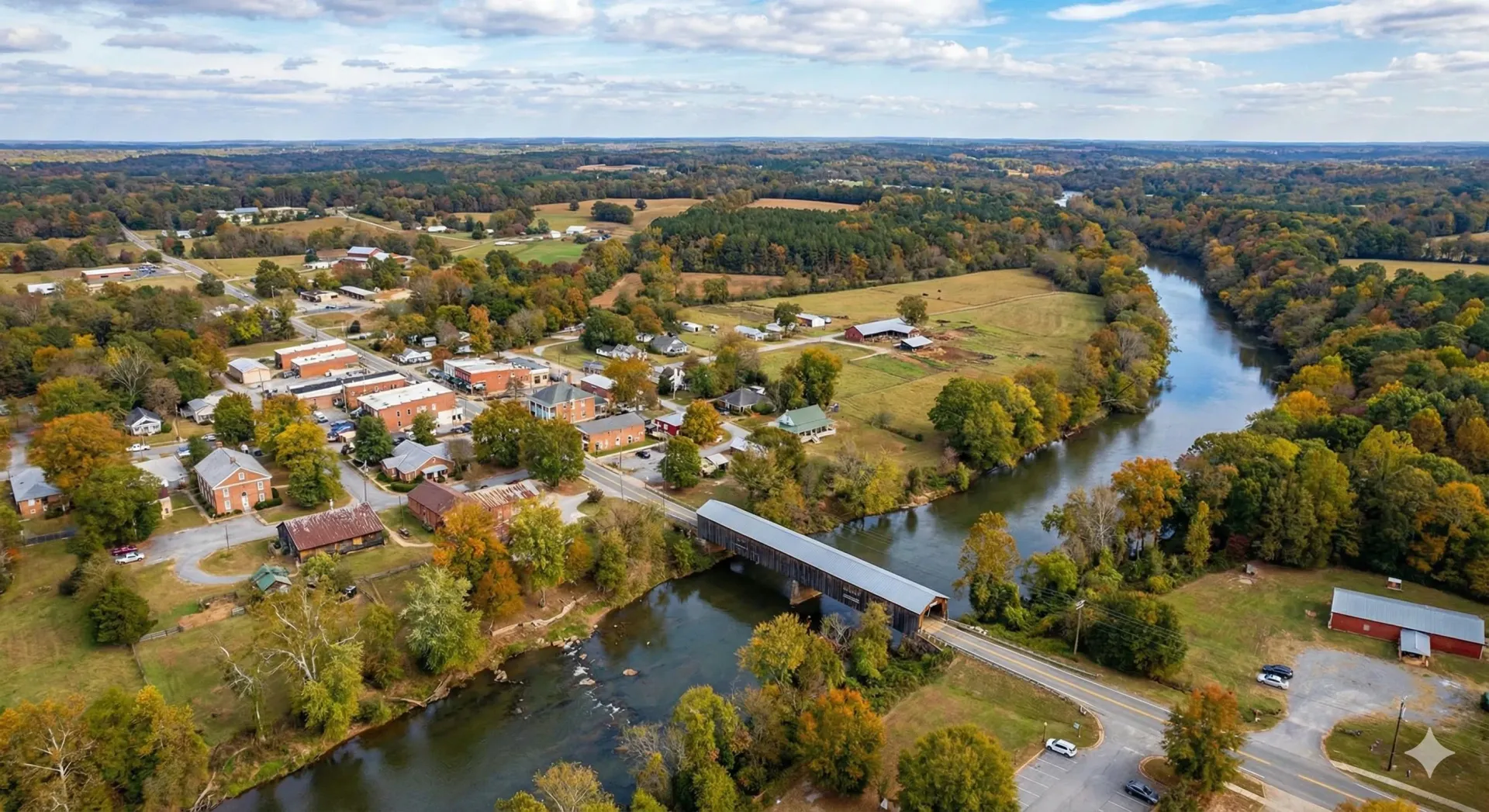 Aerial view of a town with a covered bridge over a river, surrounded by trees with autumn foliage.
