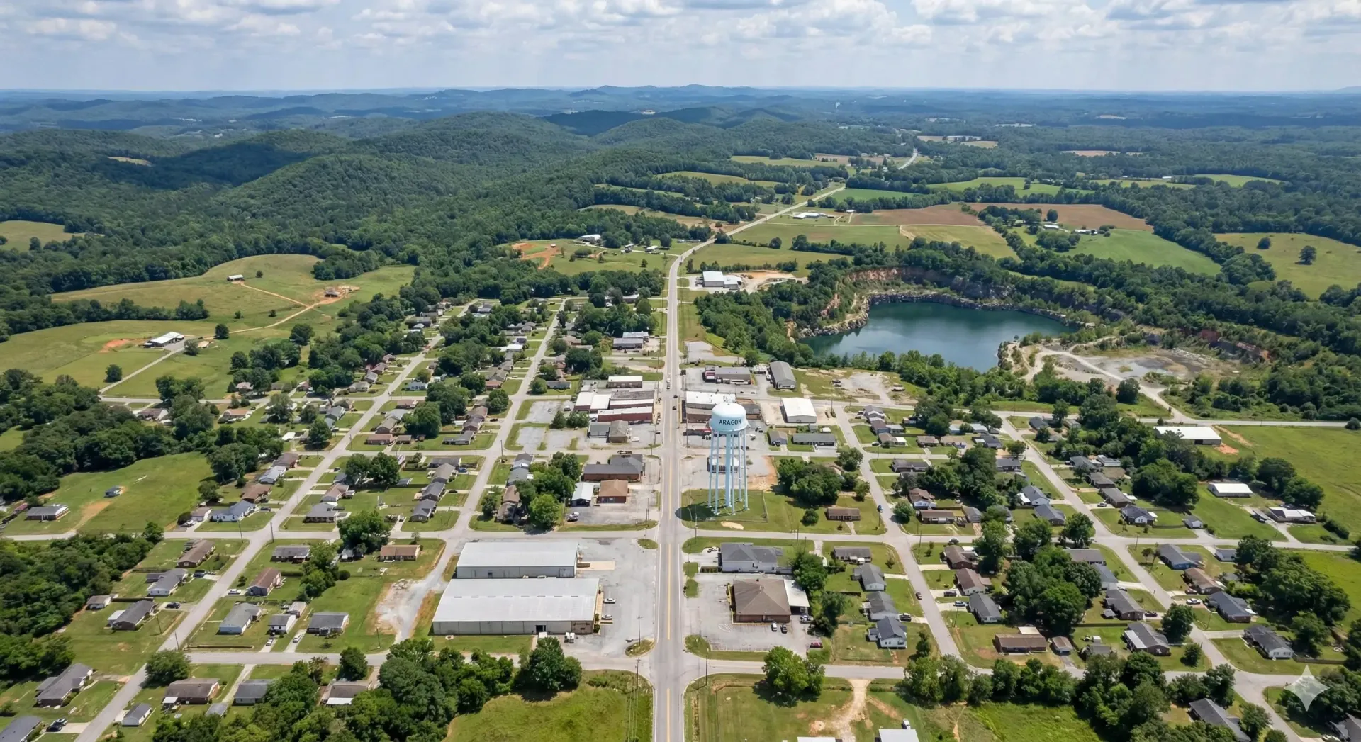 Aerial view of a small town with roads, houses, businesses, a lake, and surrounding green trees.