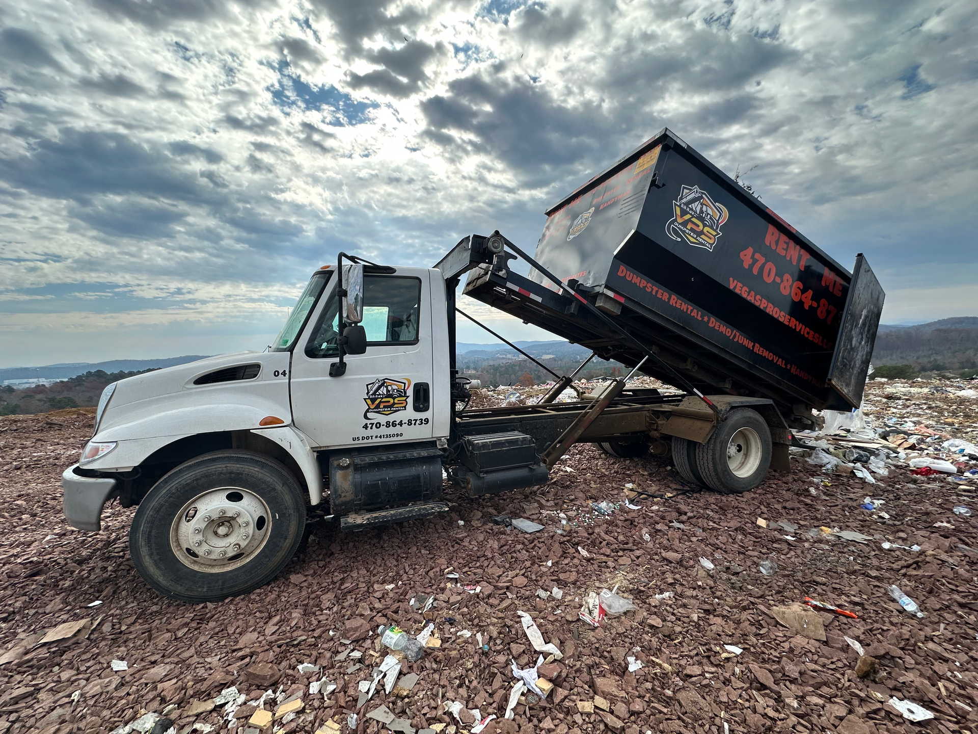 White truck with raised black dumpster on a trash-filled hillside under a cloudy sky.