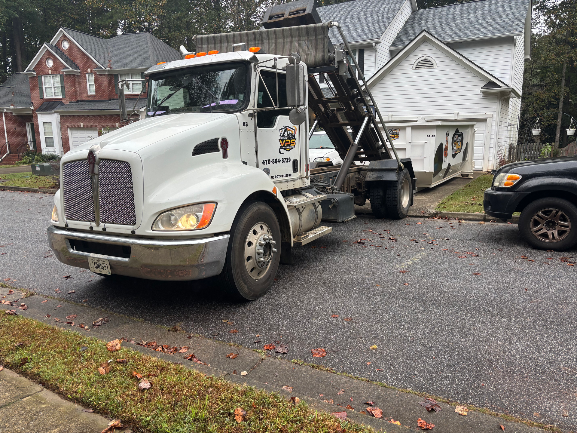 White dump truck parked in front of a house. The truck has an attached container. Asphalt road.