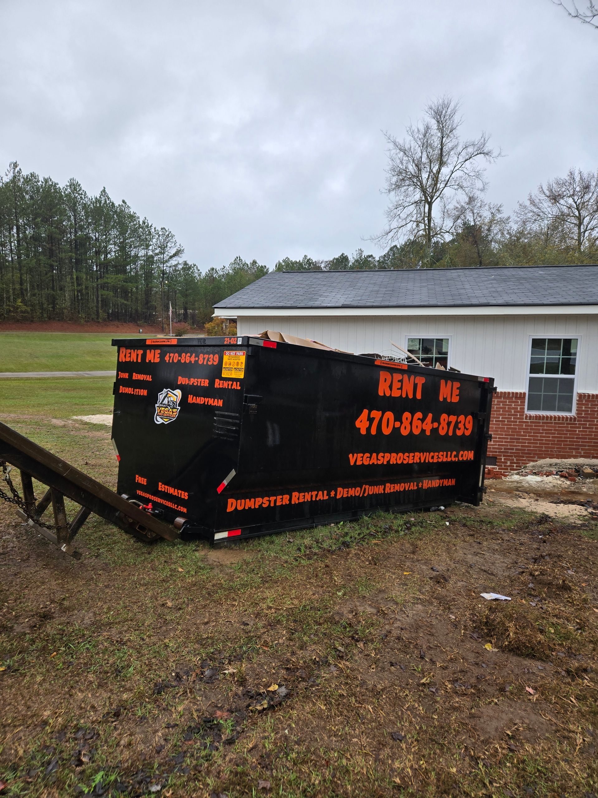 Black dumpster on a grassy yard next to a white building;