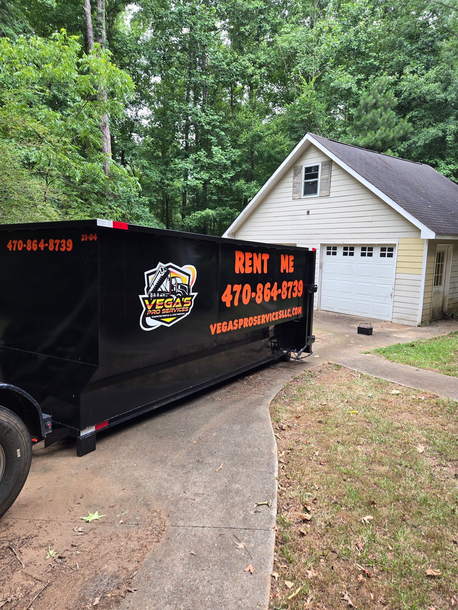 Black dumpster parked on a driveway in front of a small house.