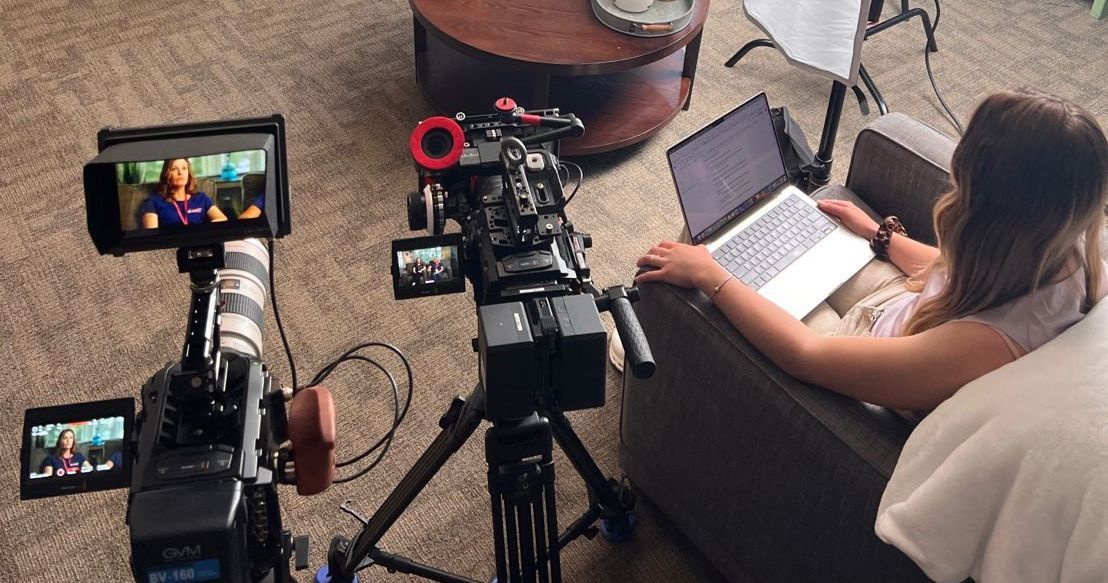 A woman is sitting on a couch with a laptop in front of a camera.