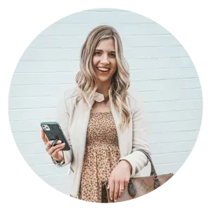 A woman is standing in front of a white wall holding a cell phone and a purse.