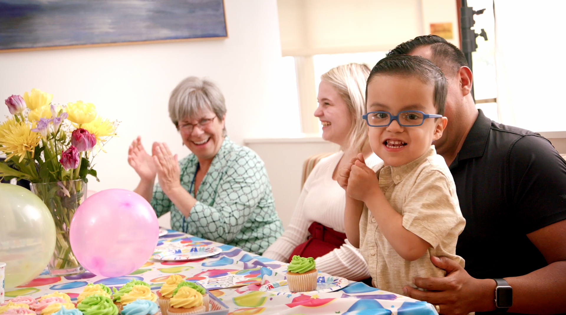 A family is sitting at a table with cupcakes and balloons at a birthday party.
