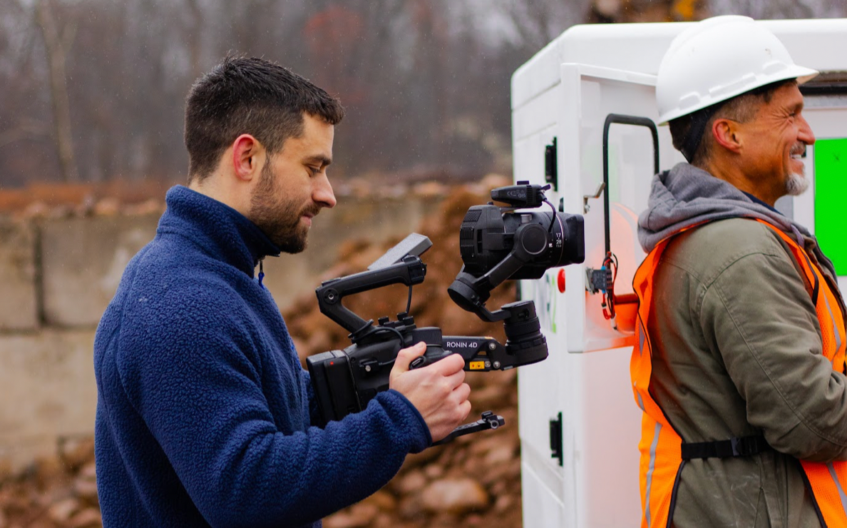 A man is holding a camera next to a man in an orange vest.