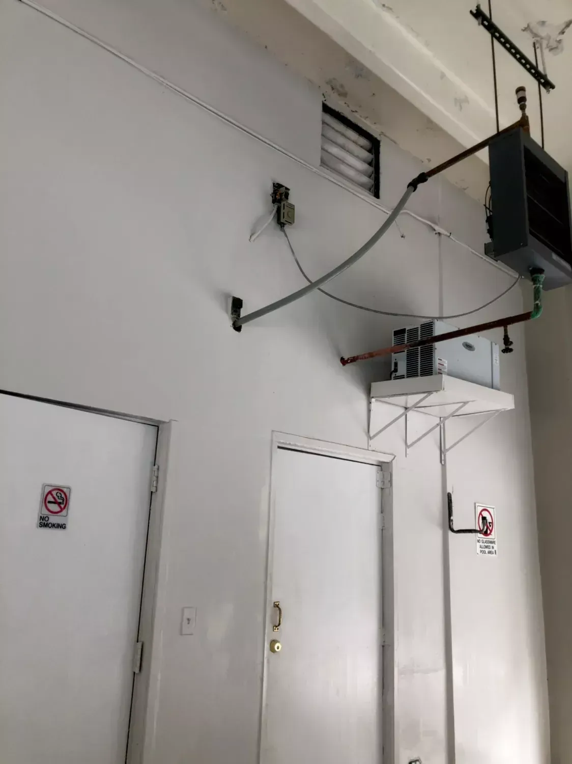White industrial hallway with doors and overhead pipes, vents, and electrical fixtures on a tall wall