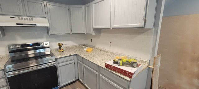 Small white kitchen with gray cabinets, stainless stove, and countertop clutter near a sink