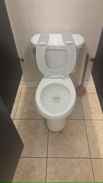 Open public restroom toilet with raised seat, beige tile floor, and toilet brush beside it.