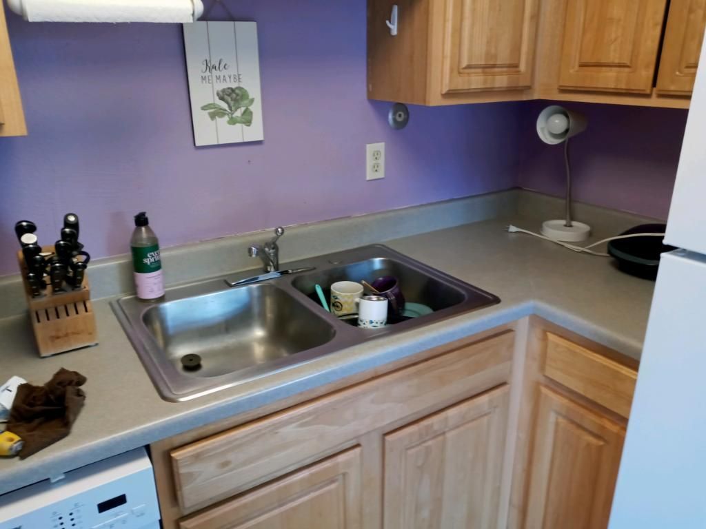 Kitchen sink and counter with wooden cabinets, lavender walls, soap bottles, and dishes by a double basin sink.