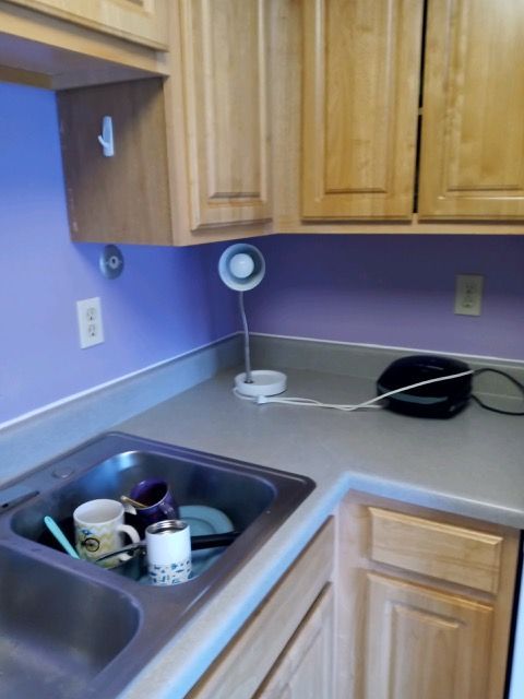 Kitchen counter and sink with dishes, a small fan, and wooden cabinets against a purple wall.