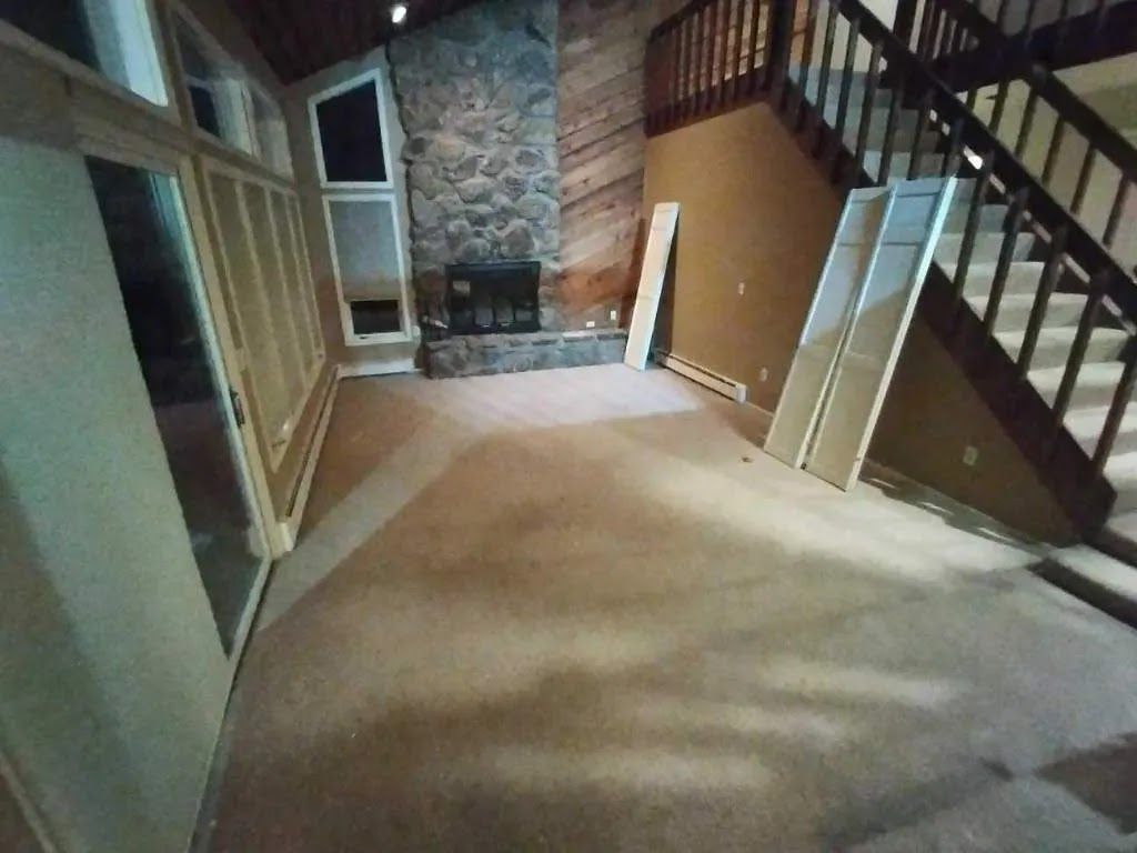 Empty living room with carpet, stone fireplace, staircase, and large sliding glass doors.