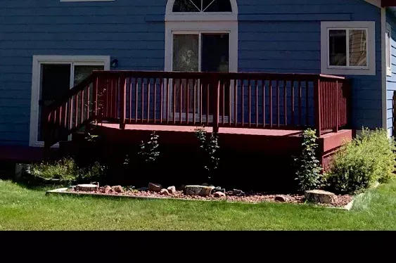 Red wooden deck attached to a blue house with white-trimmed windows and green shrubs below.