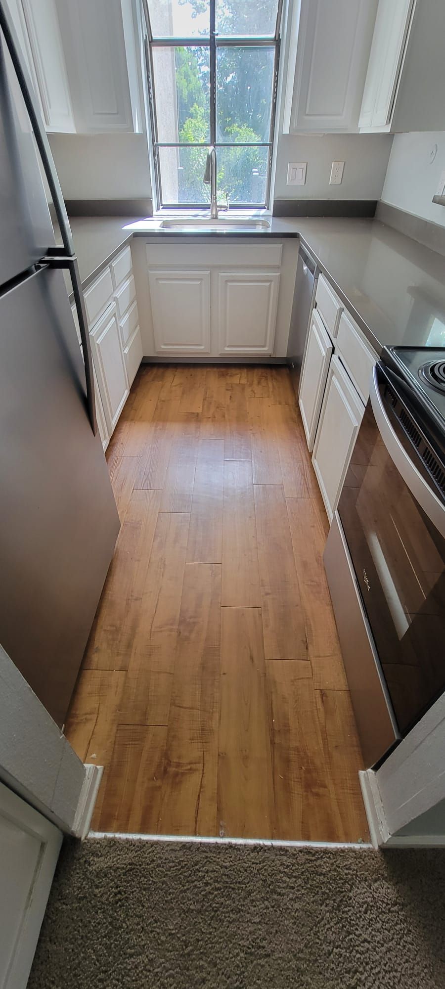 Narrow kitchen with wood floor, white cabinets, gray countertops, window, and built-in oven.