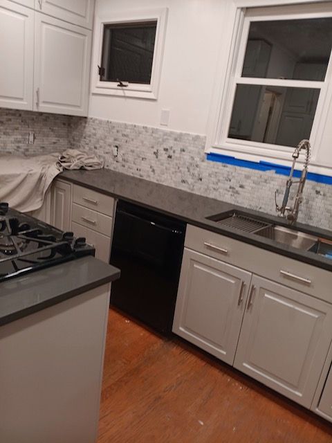 White kitchen with gray backsplash, black dishwasher, gas stove, sink, and countertop under a window.