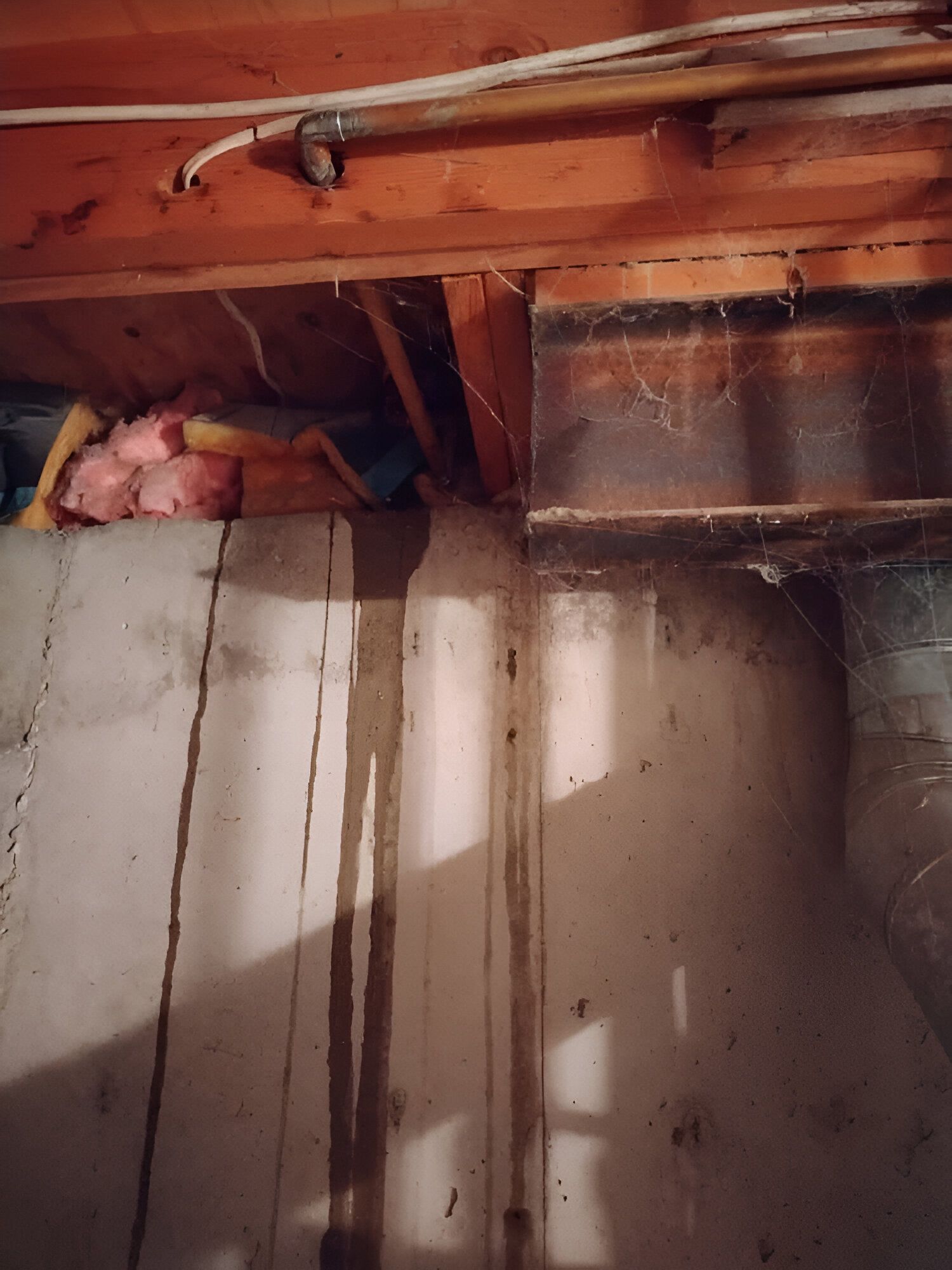 Basement wall with water stains, exposed wooden beams, insulation, and a metal duct near the ceiling