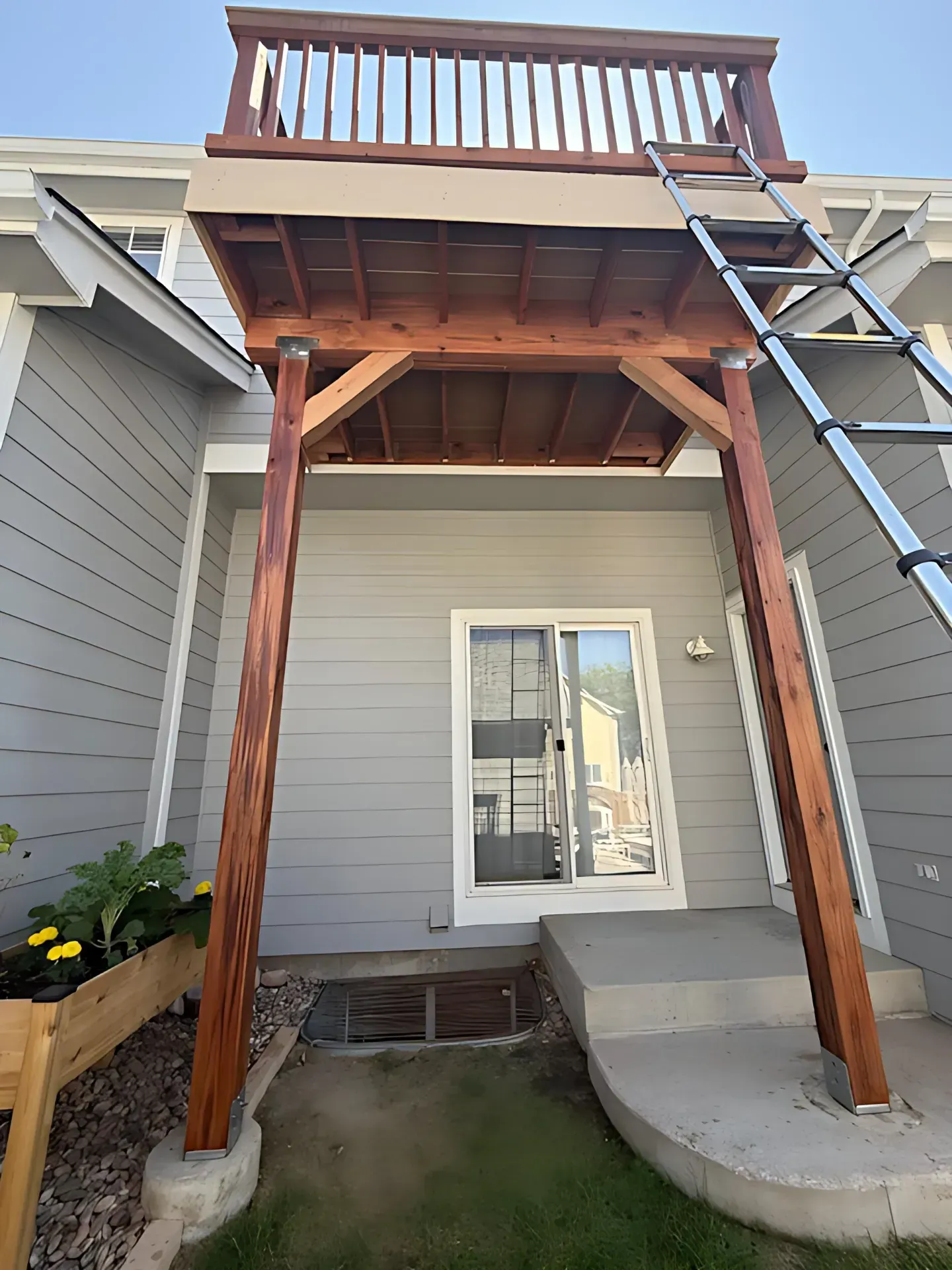 Wooden deck balcony above a small patio beside a gray house, with a ladder leaning on the right.