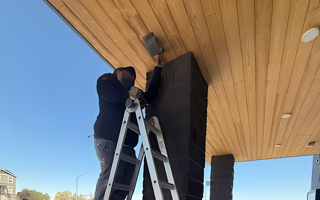 Person on a ladder installing a light under a wood-paneled overhang