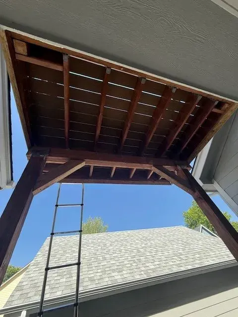 Open attic hatch with ladder, showing roof framing and blue sky above a house.