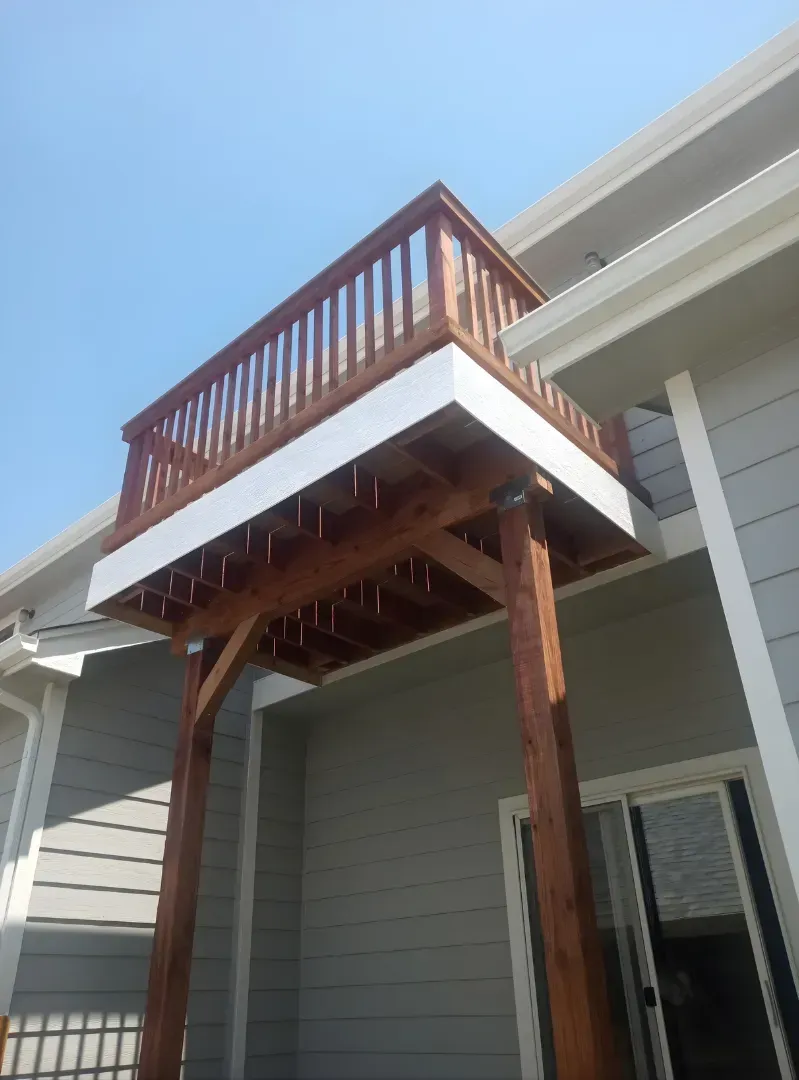 Wooden balcony deck attached to a gray house with white trim under a blue sky