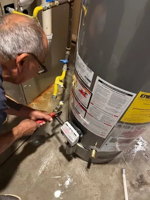 Technician inspecting a water heater, using a tool near gas pipes and a control unit in a utility room
