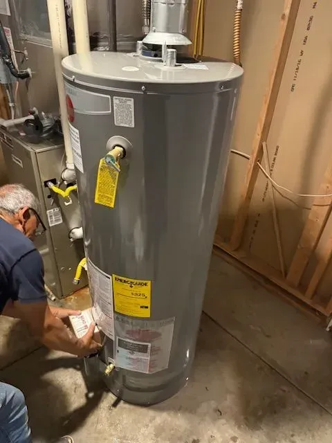 Technician inspecting a gray water heater in a utility room, with exposed pipes and construction framing nearby