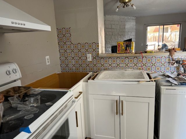Compact kitchen with white cabinets, patterned tile backsplash, stove, sink, and open cabinet under the counter