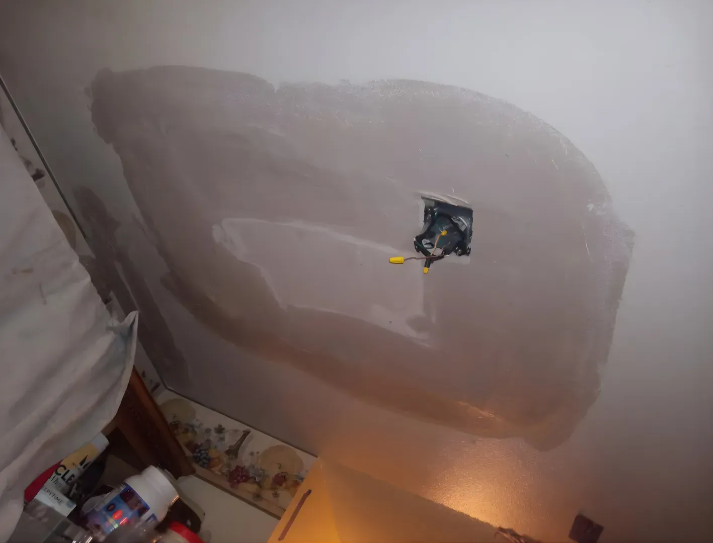 Ceiling with a large patched drywall area and exposed electrical box above a lit lamp.