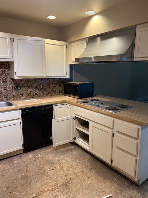 Empty kitchen with white cabinets, black dishwasher, beige tile floor, and stainless range hood under recessed lights