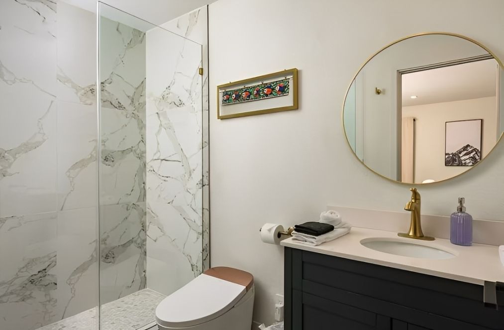 Modern bathroom with marble shower, white toilet, vanity sink, and round gold-framed mirror