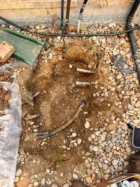 Excavated dirt hole with exposed pipes and stones in a yard beside a wall