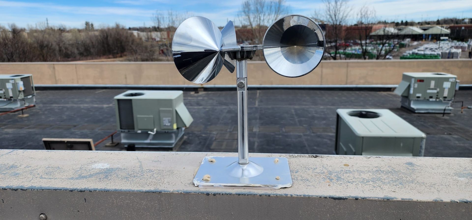 Two silver rooftop sirens on a metal pole with boxes and a low wall in the background