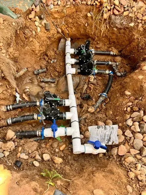 Exposed underground PVC pipe manifold with black valves in a dirt trench, one blue valve visible.