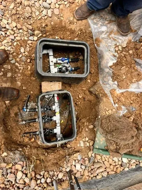 Two open utility boxes in rocky ground with exposed wires and valves, workers' boots nearby
