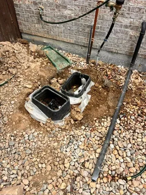 Two open utility boxes in gravel beside a brick wall, with pipes and a green cover nearby