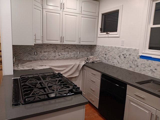 Modern kitchen with white cabinets, gray countertops, black stove, and tiled backsplash; countertop being repaired